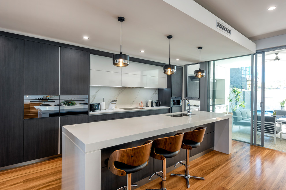 Photo of a contemporary galley kitchen in Perth with flat-panel cabinets, grey cabinets, white splashback, stainless steel appliances, medium hardwood flooring, an island and brown floors.