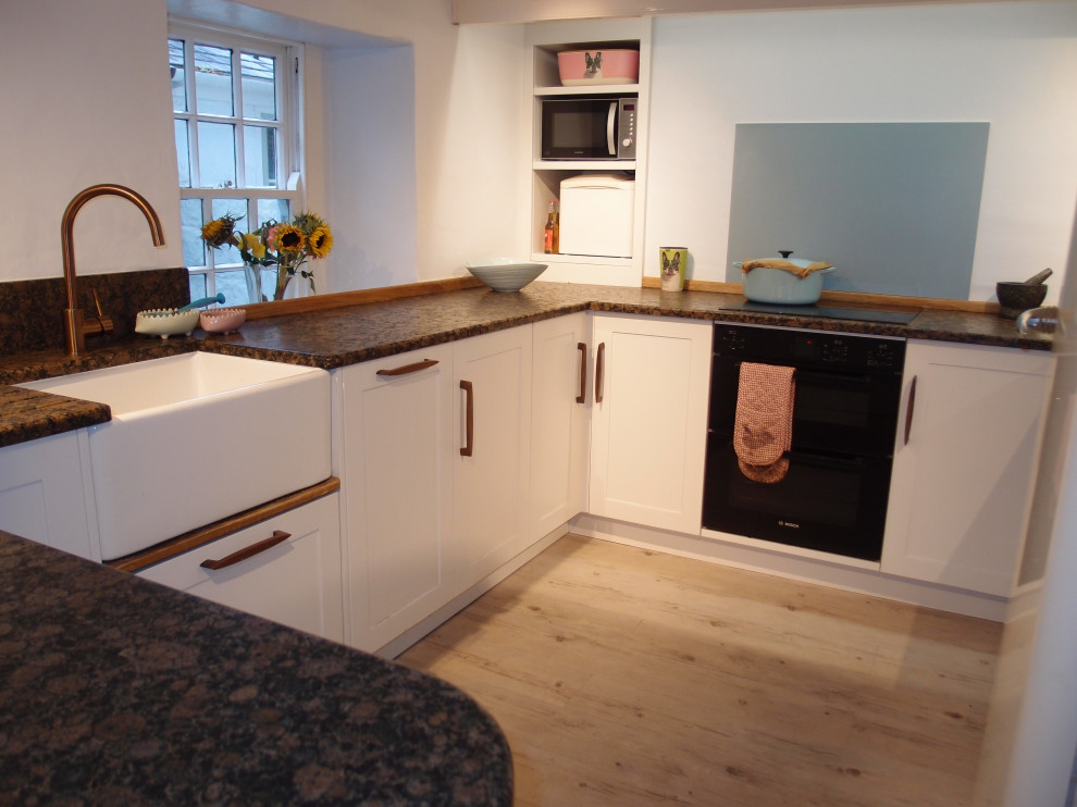 Photo of a medium sized farmhouse u-shaped enclosed kitchen in Cornwall with shaker cabinets, white cabinets, granite worktops, blue splashback, mirror splashback, black appliances, painted wood flooring, no island, white floors, brown worktops and a belfast sink.
