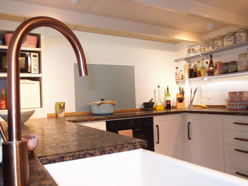 Photo of a medium sized rural u-shaped enclosed kitchen in Cornwall with a belfast sink, shaker cabinets, white cabinets, granite worktops, mirror splashback, black appliances, painted wood flooring, no island, white floors and brown worktops.