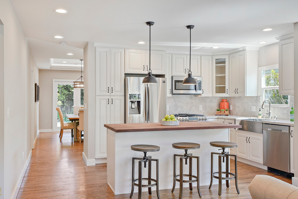 Bild på ett lantligt brun brunt l-kök, med luckor med upphöjd panel, vita skåp, träbänkskiva, grått stänkskydd, rostfria vitvaror, mellanmörkt trägolv, flera köksöar och brunt golv