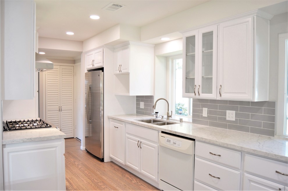 Example of a classic galley medium tone wood floor and gray floor eat-in kitchen design in Los Angeles with an undermount sink, raised-panel cabinets, white cabinets, quartz countertops, gray backsplash, ceramic backsplash, white appliances, a peninsula and gray countertops