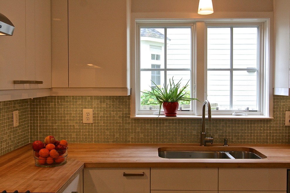 Photo of a small midcentury l-shaped kitchen/diner in DC Metro with a submerged sink, flat-panel cabinets, white cabinets, wood worktops, green splashback, glass tiled splashback, stainless steel appliances, light hardwood flooring and an island.