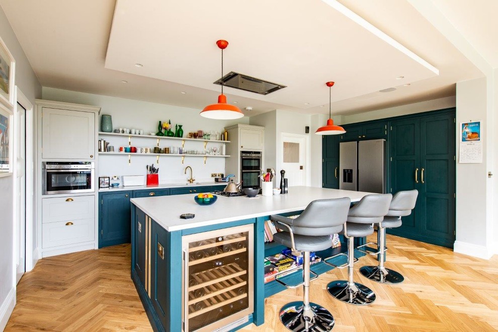 Cette photo montre une cuisine chic avec un placard à porte shaker, des portes de placard turquoises, une crédence blanche, un électroménager en acier inoxydable, parquet clair, îlot, un sol beige et un plan de travail blanc.