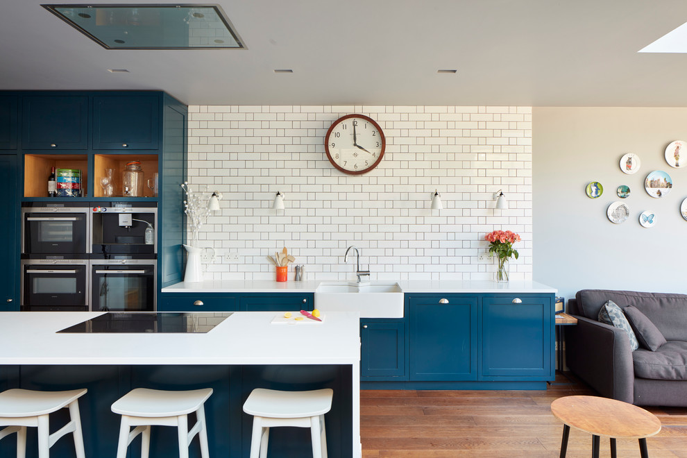 ロンドンにある高級な広いコンテンポラリースタイルのおしゃれなキッチン (エプロンフロントシンク、青いキャビネット、珪岩カウンター、白いキッチンパネル、濃色無垢フローリング、シェーカースタイル扉のキャビネット、セメントタイルのキッチンパネル、パネルと同色の調理設備、白いキッチンカウンター、三角天井) の写真