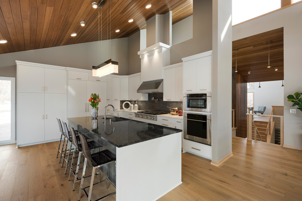 Example of a trendy l-shaped medium tone wood floor and brown floor kitchen design in Minneapolis with an undermount sink, flat-panel cabinets, white cabinets, stainless steel appliances, an island and black countertops