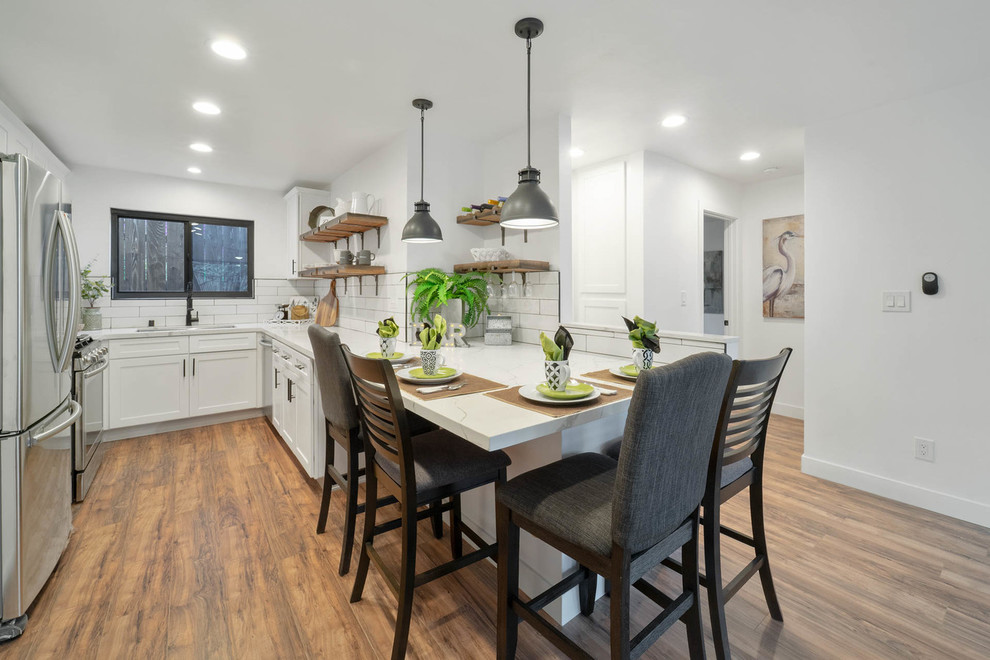 Example of a small cottage galley light wood floor and brown floor open concept kitchen design in San Diego with a double-bowl sink, flat-panel cabinets, white cabinets, quartz countertops, white backsplash, ceramic backsplash, stainless steel appliances, an island and white countertops