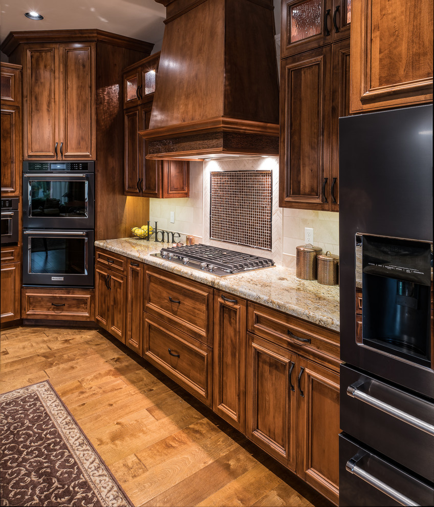 Example of a mid-sized mountain style l-shaped medium tone wood floor and brown floor open concept kitchen design in Minneapolis with a single-bowl sink, raised-panel cabinets, medium tone wood cabinets, granite countertops, beige backsplash, stone tile backsplash, black appliances and an island