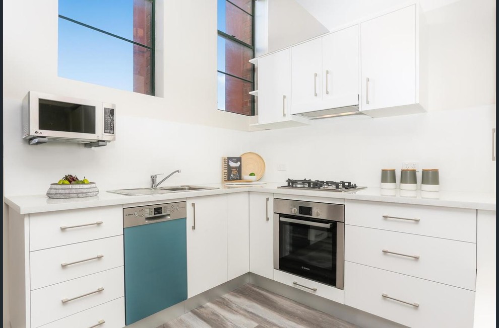 Design ideas for a small contemporary l-shaped open plan kitchen in Sydney with a single-bowl sink, flat-panel cabinets, white cabinets, engineered stone countertops, white splashback, ceramic splashback, stainless steel appliances, laminate floors, no island, grey floors and white worktops.
