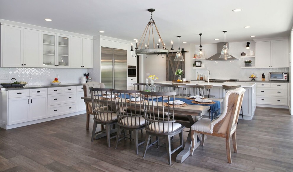 Photo of a large rural l-shaped kitchen/diner in Orange County with a belfast sink, shaker cabinets, white cabinets, engineered stone countertops, white splashback, ceramic splashback, stainless steel appliances, medium hardwood flooring, an island, grey floors and grey worktops.