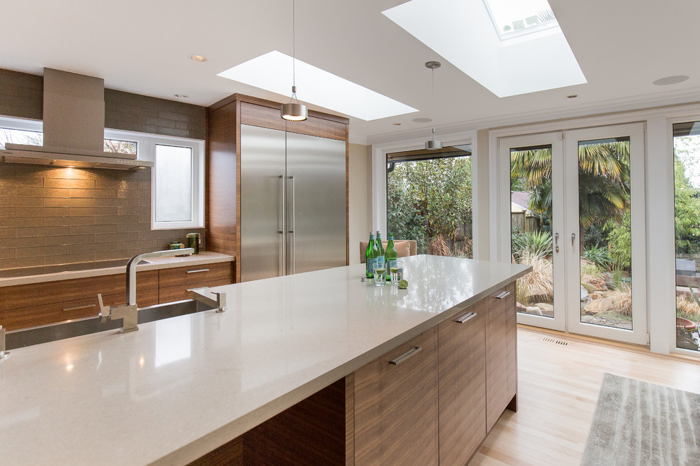This is an example of a medium sized contemporary l-shaped open plan kitchen in Vancouver with a built-in sink, flat-panel cabinets, brown cabinets, grey splashback, glass tiled splashback, stainless steel appliances, light hardwood flooring and an island.