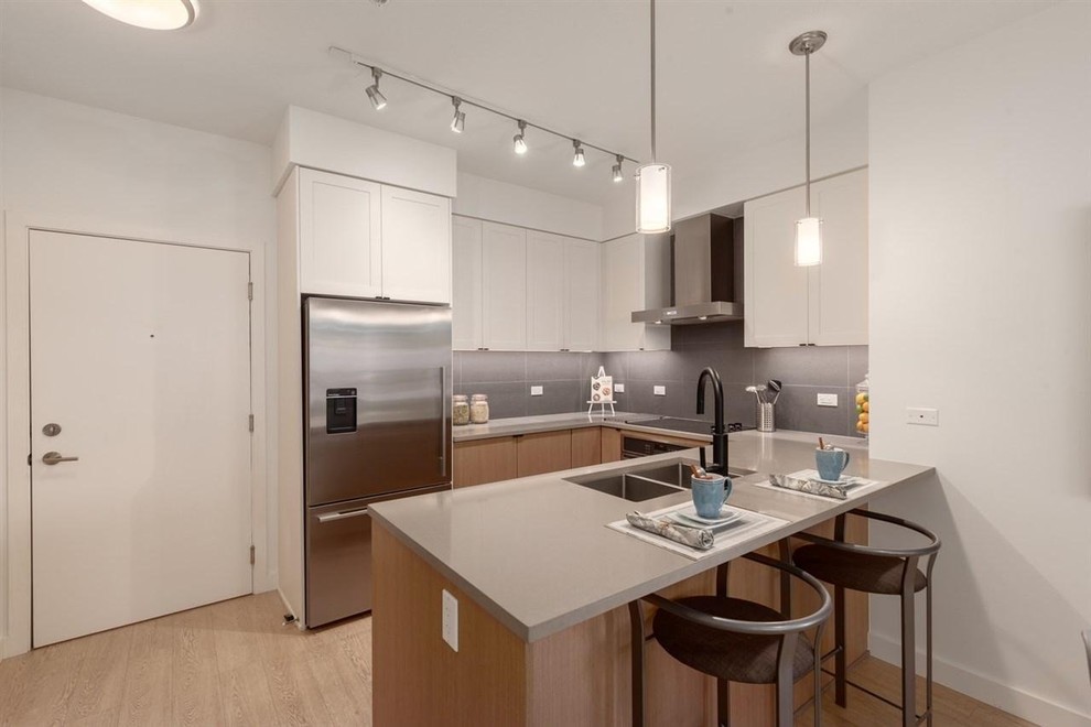 Small contemporary u-shaped open plan kitchen in Vancouver with a double-bowl sink, recessed-panel cabinets, white cabinets, composite countertops, grey splashback, porcelain splashback, stainless steel appliances, medium hardwood flooring, a breakfast bar, brown floors and grey worktops.