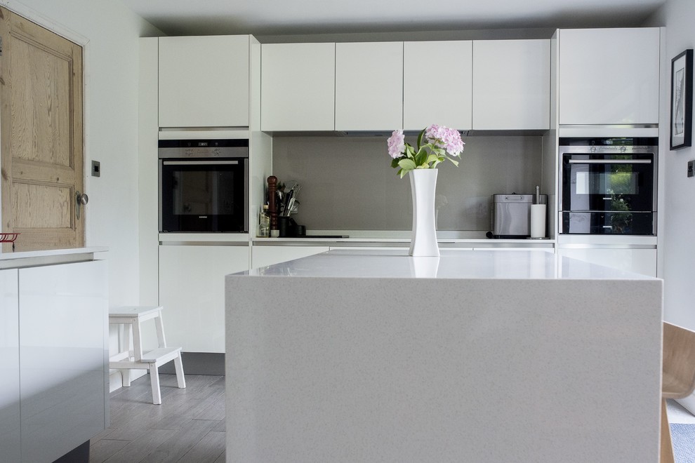 Inredning av ett modernt mellanstort grå grått kök, med en enkel diskho, vita skåp, bänkskiva i kvartsit, grått stänkskydd, glaspanel som stänkskydd, svarta vitvaror, klinkergolv i porslin, en köksö och grått golv
