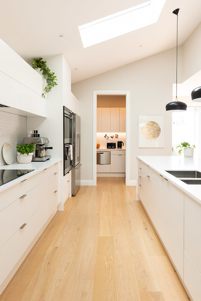 Kitchen - contemporary galley light wood floor and beige floor kitchen idea in Auckland with an undermount sink, white cabinets, black appliances, white countertops, recessed-panel cabinets, solid surface countertops, white backsplash and ceramic backsplash