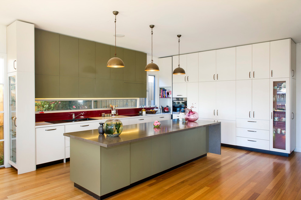 Photo of a large classic l-shaped open plan kitchen in Melbourne with flat-panel cabinets, green cabinets, red splashback, light hardwood flooring, an island, a double-bowl sink, engineered stone countertops, glass sheet splashback, stainless steel appliances, brown floors and brown worktops.