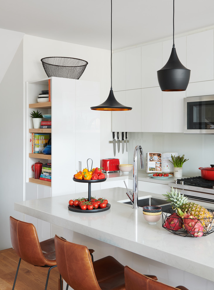 This is an example of a contemporary kitchen in Toronto with a submerged sink, flat-panel cabinets, white cabinets, white splashback, glass sheet splashback, stainless steel appliances, medium hardwood flooring, an island, brown floors and white worktops.