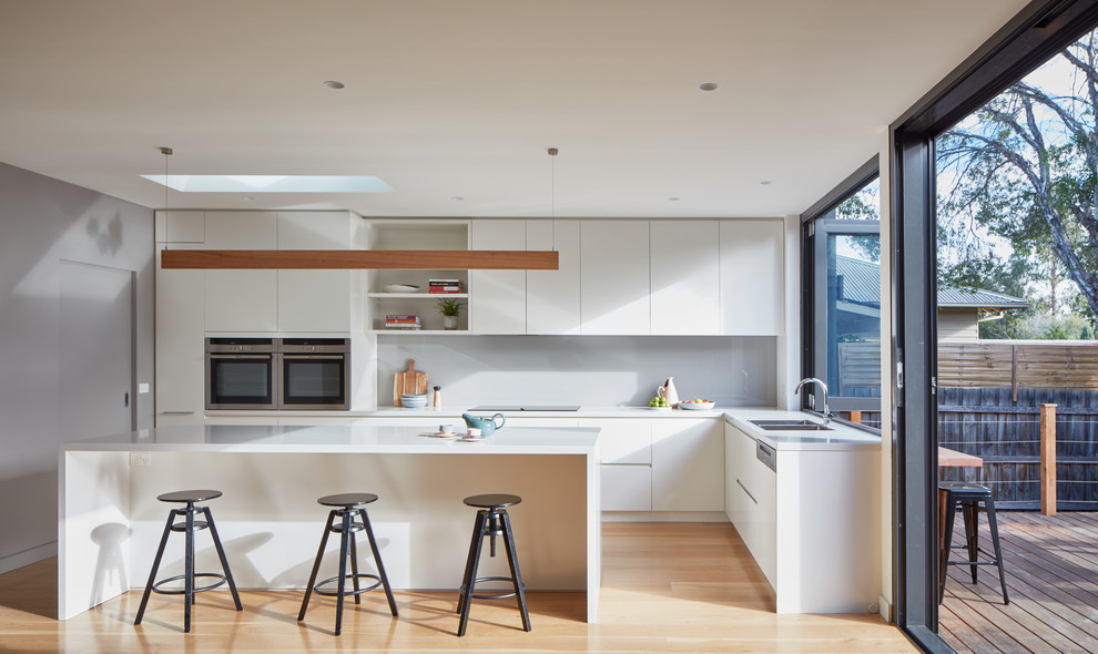Moderne Wohnküche in L-Form mit Doppelwaschbecken, Küchenrückwand in Grau, Glasrückwand, hellem Holzboden, Kücheninsel und Küchengeräten aus Edelstahl in Melbourne