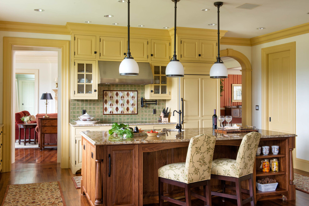 Inredning av ett klassiskt stort, avskilt l-kök, med en undermonterad diskho, luckor med upphöjd panel, gula skåp, flerfärgad stänkskydd, integrerade vitvaror, mellanmörkt trägolv, en köksö, granitbänkskiva och stänkskydd i keramik
