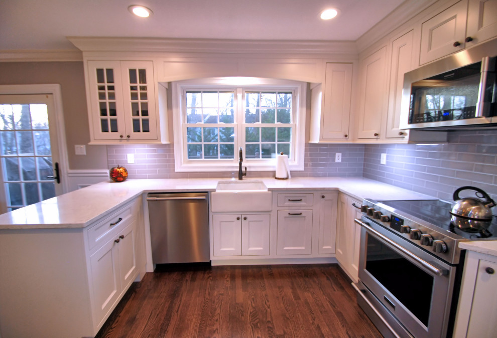 Inredning av ett klassiskt mellanstort vit vitt kök, med en rustik diskho, luckor med profilerade fronter, vita skåp, bänkskiva i kvarts, blått stänkskydd, stänkskydd i glaskakel, rostfria vitvaror, mellanmörkt trägolv, en halv köksö och brunt golv