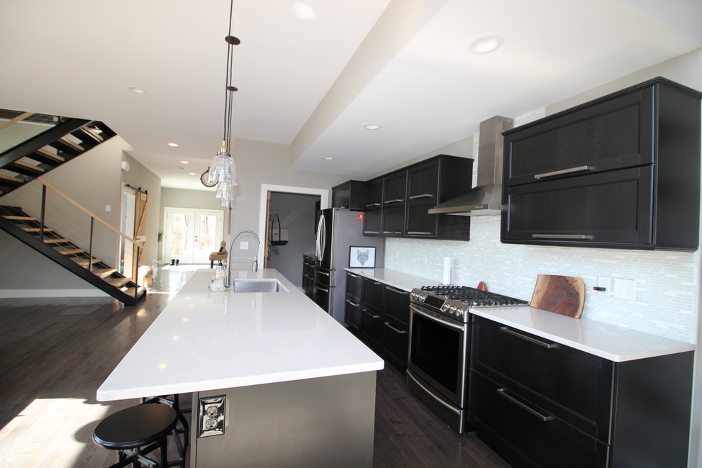 Photo of a large modern single-wall open plan kitchen in Cincinnati with a submerged sink, recessed-panel cabinets, black cabinets, composite countertops, white splashback, matchstick tiled splashback, stainless steel appliances, dark hardwood flooring and an island.