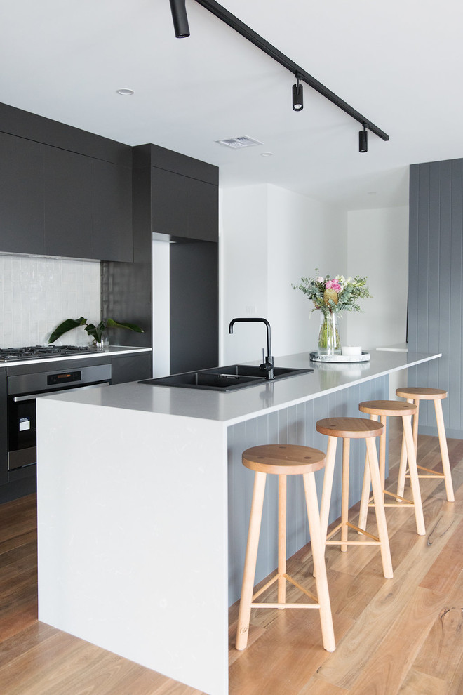 This is an example of a medium sized modern galley open plan kitchen in Canberra - Queanbeyan with a double-bowl sink, black cabinets, engineered stone countertops, grey splashback, metro tiled splashback, black appliances, medium hardwood flooring, an island and brown floors.