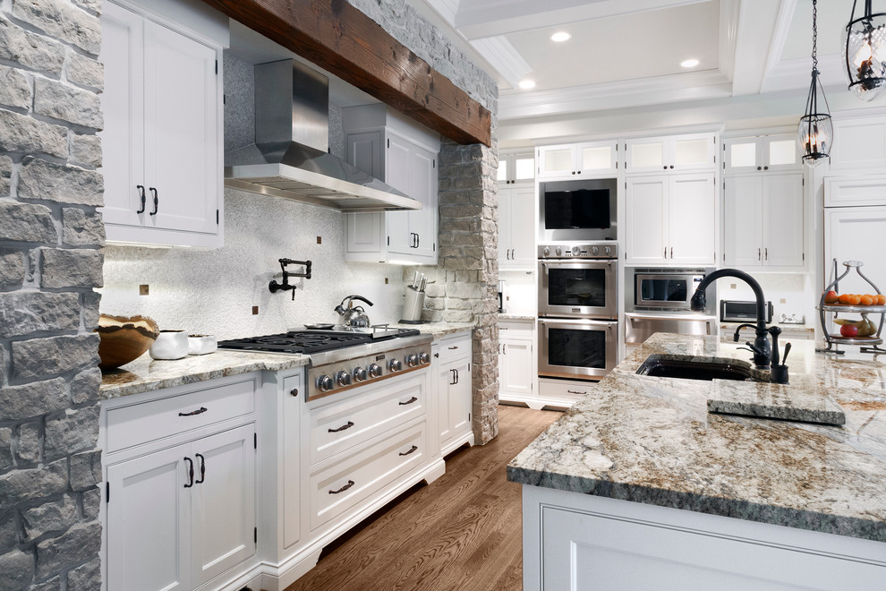 Cette photo montre une cuisine chic avec un évier encastré, un placard à porte affleurante, des portes de placard blanches, un plan de travail en granite, une crédence grise et un électroménager en acier inoxydable.