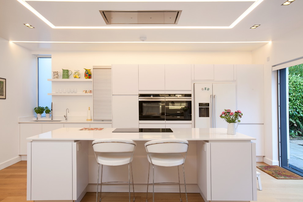 Medium sized contemporary single-wall open plan kitchen in Other with flat-panel cabinets, white cabinets, laminate countertops, white splashback, integrated appliances, light hardwood flooring, an island, beige floors, white worktops and a submerged sink.