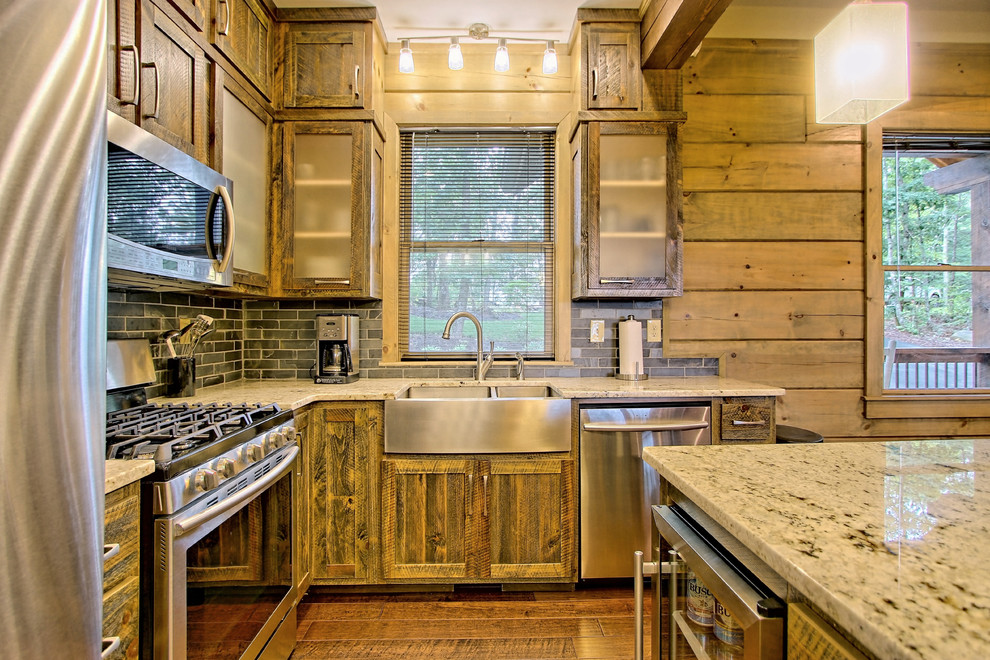 Small mountain style l-shaped medium tone wood floor and brown floor open concept kitchen photo in Other with a farmhouse sink, recessed-panel cabinets, distressed cabinets, granite countertops, gray backsplash, stone tile backsplash, stainless steel appliances and an island