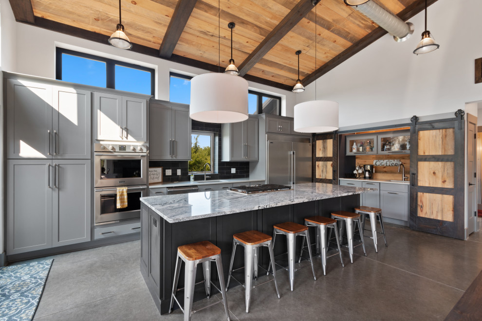 Large industrial l-shaped open plan kitchen in Milwaukee with a submerged sink, shaker cabinets, grey cabinets, granite worktops, grey splashback, metro tiled splashback, stainless steel appliances, concrete flooring, an island, brown floors, multicoloured worktops and a wood ceiling.