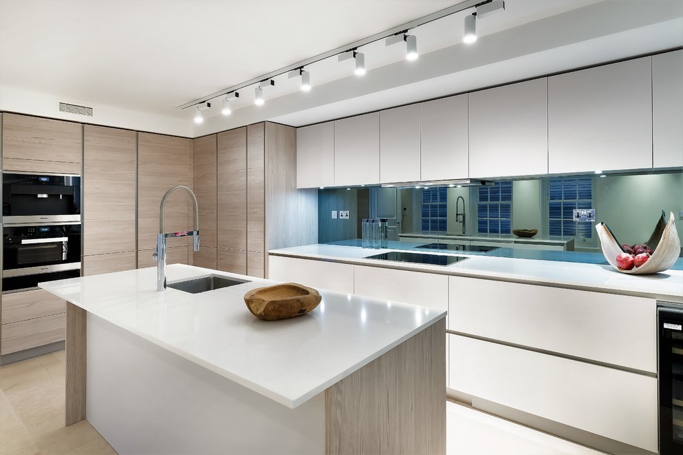This is an example of a large contemporary l-shaped kitchen/diner in London with a submerged sink, flat-panel cabinets, white cabinets, mirror splashback, black appliances, an island, beige floors, white worktops, composite countertops, metallic splashback and limestone flooring.