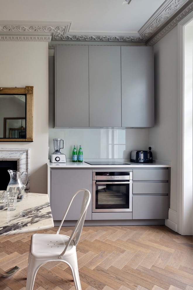 Medium sized eclectic single-wall kitchen/diner in London with a single-bowl sink, flat-panel cabinets, grey cabinets, composite countertops, grey splashback, glass sheet splashback, stainless steel appliances, medium hardwood flooring, no island, brown floors and white worktops.