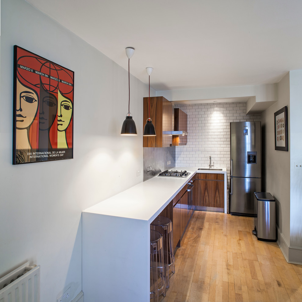 Contemporary l-shaped enclosed kitchen in London with a submerged sink, flat-panel cabinets, white splashback, stainless steel appliances, metro tiled splashback, medium wood cabinets and white worktops.
