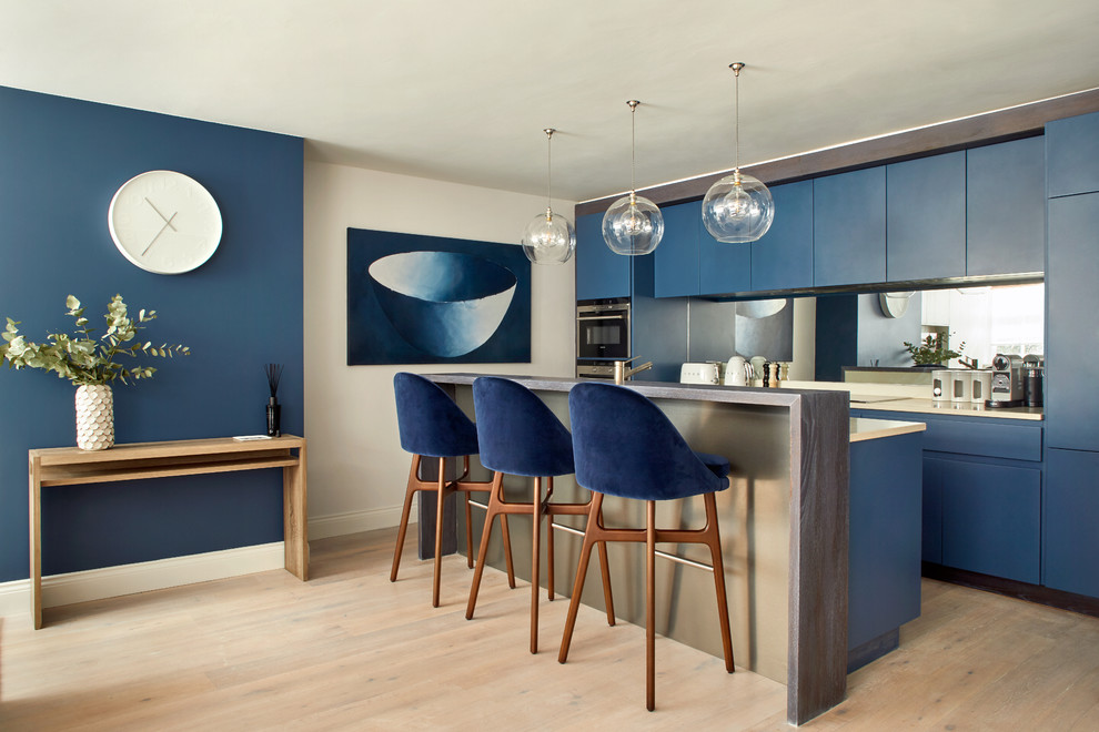 Photo of a contemporary galley kitchen in London with flat-panel cabinets, blue cabinets, composite countertops, mirror splashback, light hardwood flooring, an island, beige worktops and beige floors.