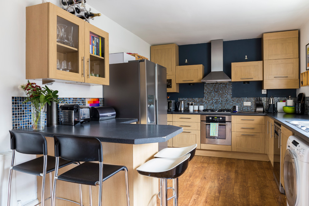 Klassische Küche in U-Form mit Schrankfronten im Shaker-Stil, hellen Holzschränken, Küchenrückwand in Blau, Küchengeräten aus Edelstahl, braunem Holzboden und Halbinsel in London