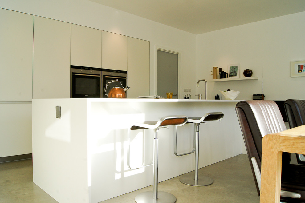 Свежая идея для дизайна: прямая кухня среднего размера в современном стиле с обеденным столом, врезной мойкой, плоскими фасадами, белыми фасадами, стеклянной столешницей, черной техникой, полом из сланца и островом - отличное фото интерьера