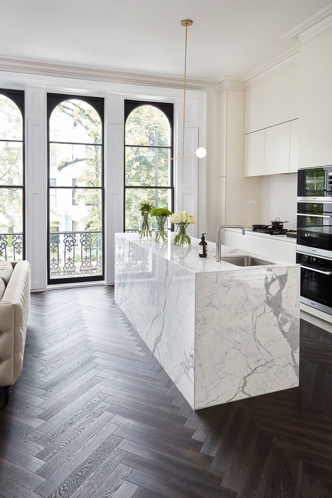 Modern inredning av ett litet vit linjärt vitt kök med öppen planlösning, med släta luckor, vita skåp, vitt stänkskydd, mörkt trägolv, en köksö, brunt golv, en undermonterad diskho och marmorbänkskiva