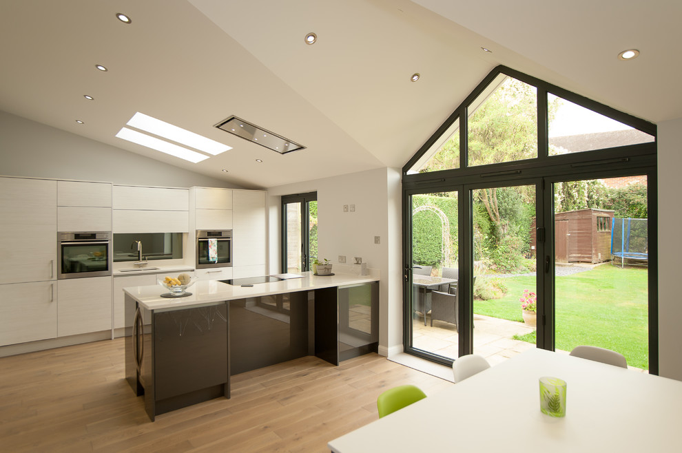 Design ideas for a medium sized modern l-shaped kitchen/diner in Hertfordshire with a built-in sink, flat-panel cabinets, white cabinets, laminate countertops, black splashback, glass sheet splashback, stainless steel appliances, light hardwood flooring, an island and brown floors.