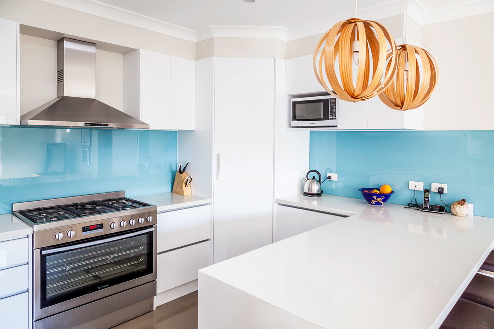 Moderne Wohnküche in L-Form mit Unterbauwaschbecken, Küchenrückwand in Blau, Glasrückwand, Küchengeräten aus Edelstahl, Keramikboden und weißen Schränken in Melbourne