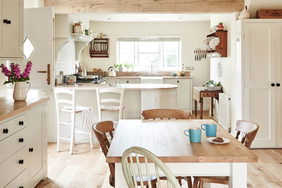 Photo of a rural l-shaped kitchen/diner in Wiltshire with a belfast sink, shaker cabinets, white cabinets, white splashback, light hardwood flooring, a breakfast bar and beige floors.