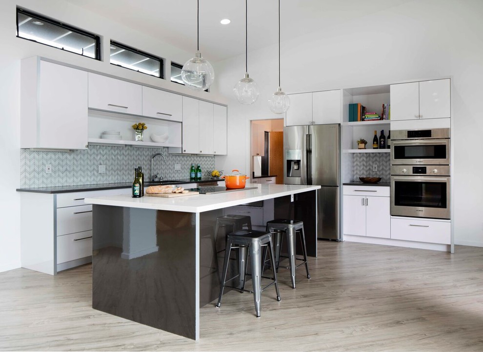 Foto på ett mellanstort funkis l-kök, med släta luckor, vita skåp, rostfria vitvaror, en köksö, en undermonterad diskho, bänkskiva i kvartsit, grått stänkskydd, stänkskydd i glaskakel och ljust trägolv