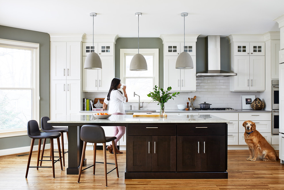 Photo of a traditional single-wall kitchen in DC Metro with a submerged sink, shaker cabinets, white cabinets, white splashback, metro tiled splashback, stainless steel appliances, medium hardwood flooring, an island, brown floors and white worktops.