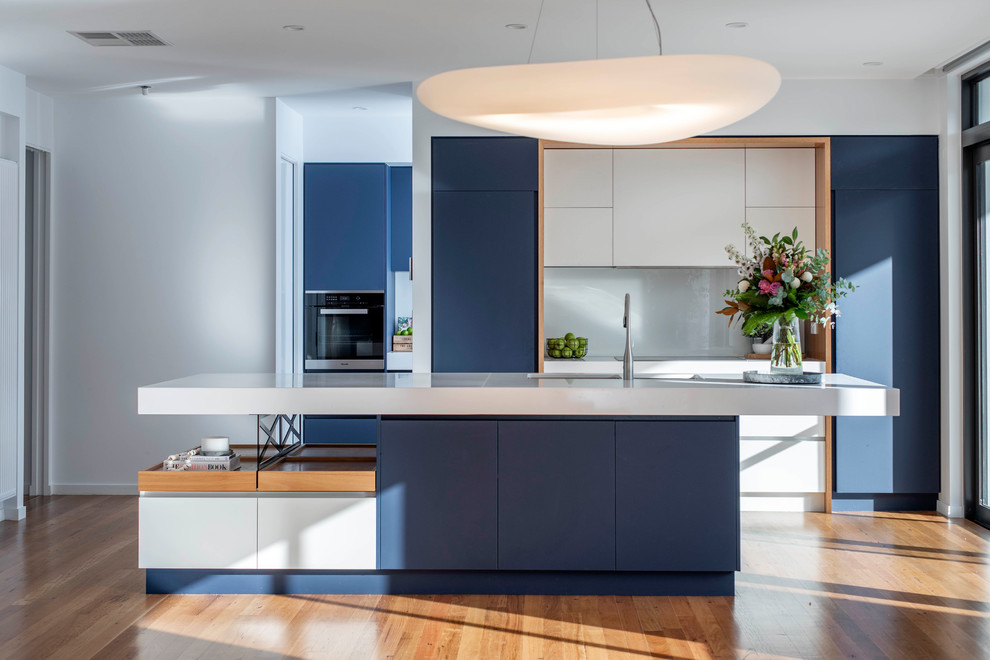 Design ideas for a contemporary galley open plan kitchen in Canberra - Queanbeyan with a submerged sink, flat-panel cabinets, blue cabinets, grey splashback, integrated appliances, medium hardwood flooring, an island and white worktops.