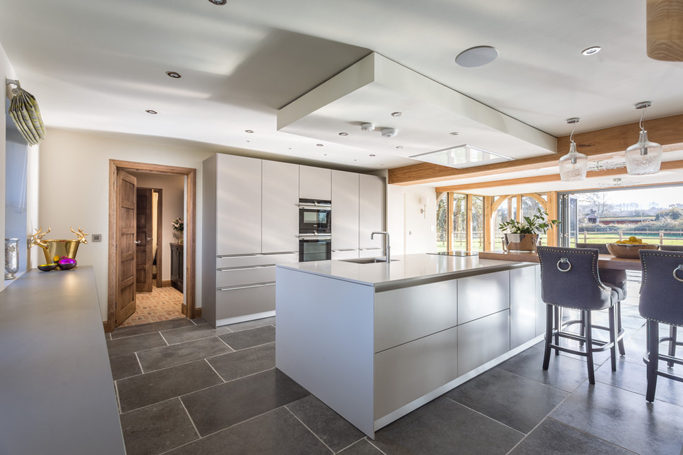 Foto på ett stort funkis linjärt kök och matrum, med en enkel diskho, släta luckor, vita skåp, bänkskiva i kvartsit, svarta vitvaror, klinkergolv i porslin och en köksö