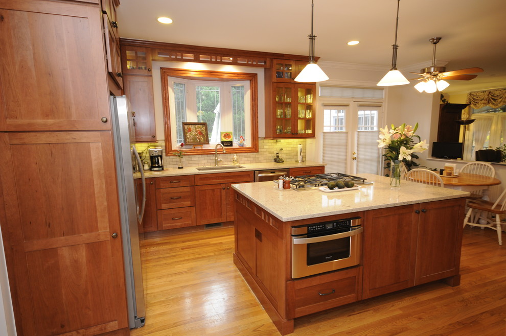 Foto på ett mellanstort amerikanskt kök, med en enkel diskho, skåp i shakerstil, skåp i mellenmörkt trä, bänkskiva i kvarts, grått stänkskydd, stänkskydd i stenkakel, rostfria vitvaror, ljust trägolv och en köksö