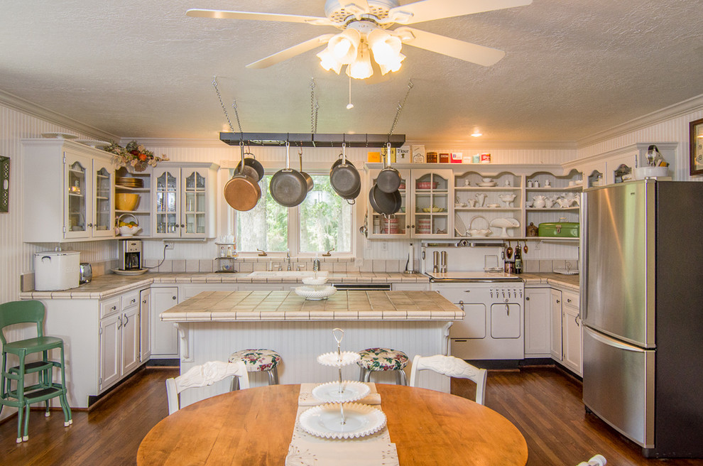 Design ideas for a large farmhouse u-shaped kitchen/diner in Houston with a double-bowl sink, glass-front cabinets, white cabinets, tile countertops, beige splashback, ceramic splashback, white appliances, medium hardwood flooring and an island.