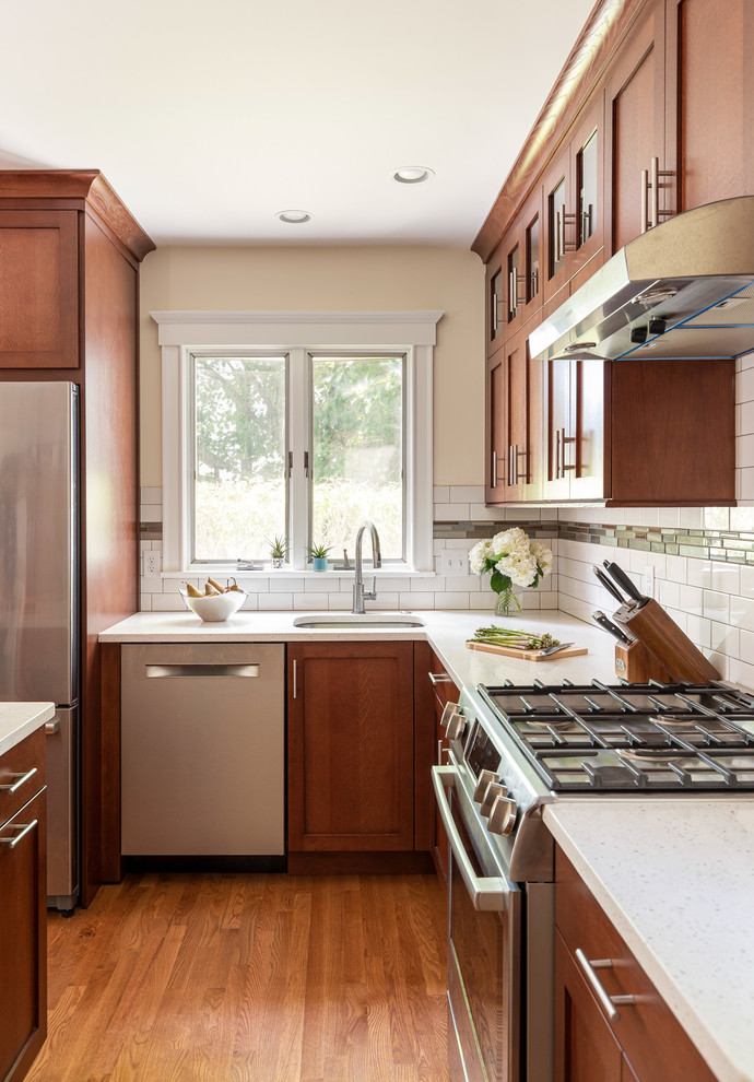 This is an example of a medium sized classic l-shaped kitchen pantry in Boston with a submerged sink, shaker cabinets, medium wood cabinets, engineered stone countertops, white splashback, metro tiled splashback, stainless steel appliances, medium hardwood flooring, an island, brown floors and white worktops.