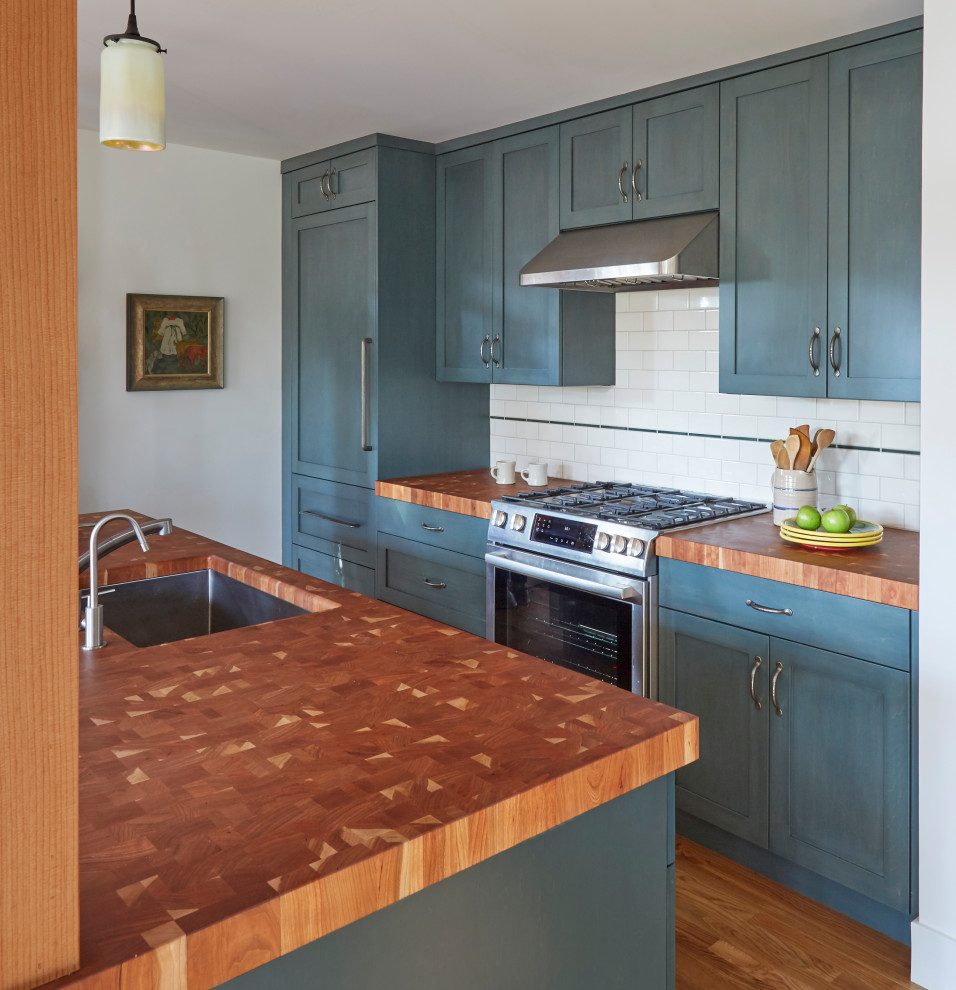 Idéer för att renovera ett mellanstort amerikanskt brun linjärt brunt kök med öppen planlösning, med en undermonterad diskho, skåp i shakerstil, blå skåp, träbänkskiva, vitt stänkskydd, stänkskydd i porslinskakel, integrerade vitvaror, mellanmörkt trägolv, en köksö och brunt golv