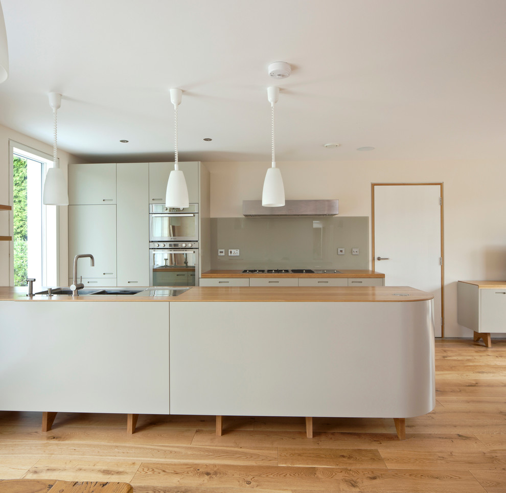 Medium sized contemporary kitchen in London with a double-bowl sink, flat-panel cabinets, grey cabinets, wood worktops, grey splashback, glass sheet splashback, stainless steel appliances, an island and light hardwood flooring.