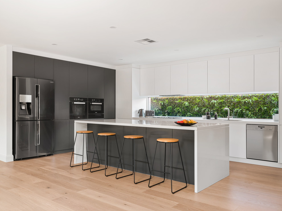 This is an example of a large contemporary l-shaped open plan kitchen in Sydney with a double-bowl sink, composite countertops, window splashback, black appliances, light hardwood flooring, an island, white worktops, flat-panel cabinets, white cabinets and beige floors.