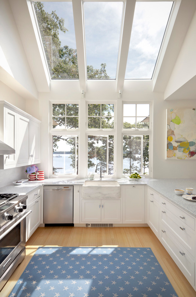 This is an example of a beach style u-shaped kitchen in Portland Maine with a belfast sink, shaker cabinets, white cabinets, stainless steel appliances and light hardwood flooring.