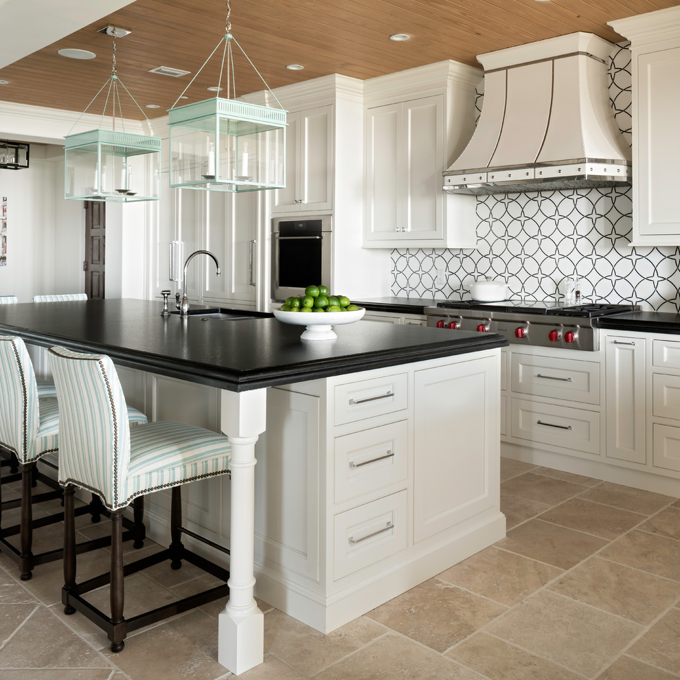 This is an example of a beach style l-shaped kitchen/diner with a submerged sink, recessed-panel cabinets, white cabinets, multi-coloured splashback, integrated appliances, an island, beige floors and black worktops.